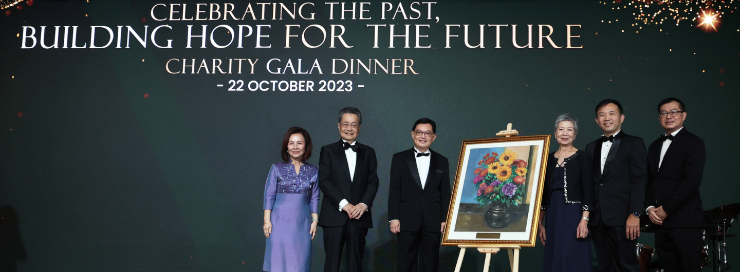 People in formal attire pose with a framed painting of flowers under event text on a dark background.